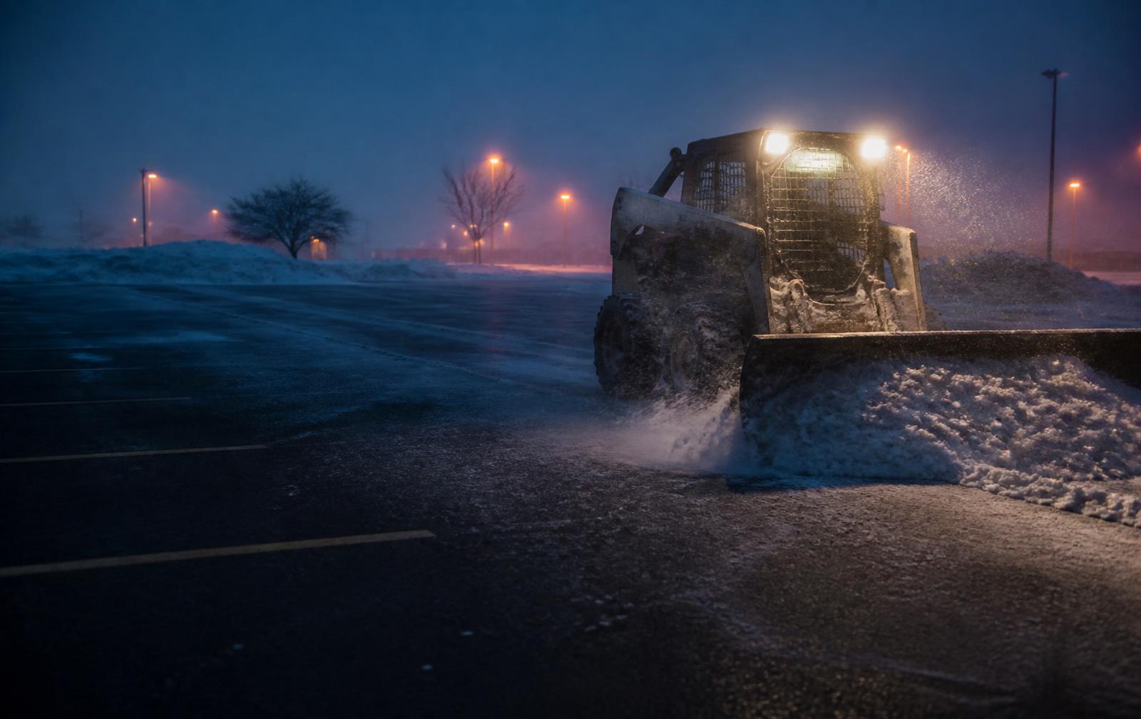 Skid steer with a snow pusher clearing a commercial parking lot before dawn with salt scattered across wet pavement