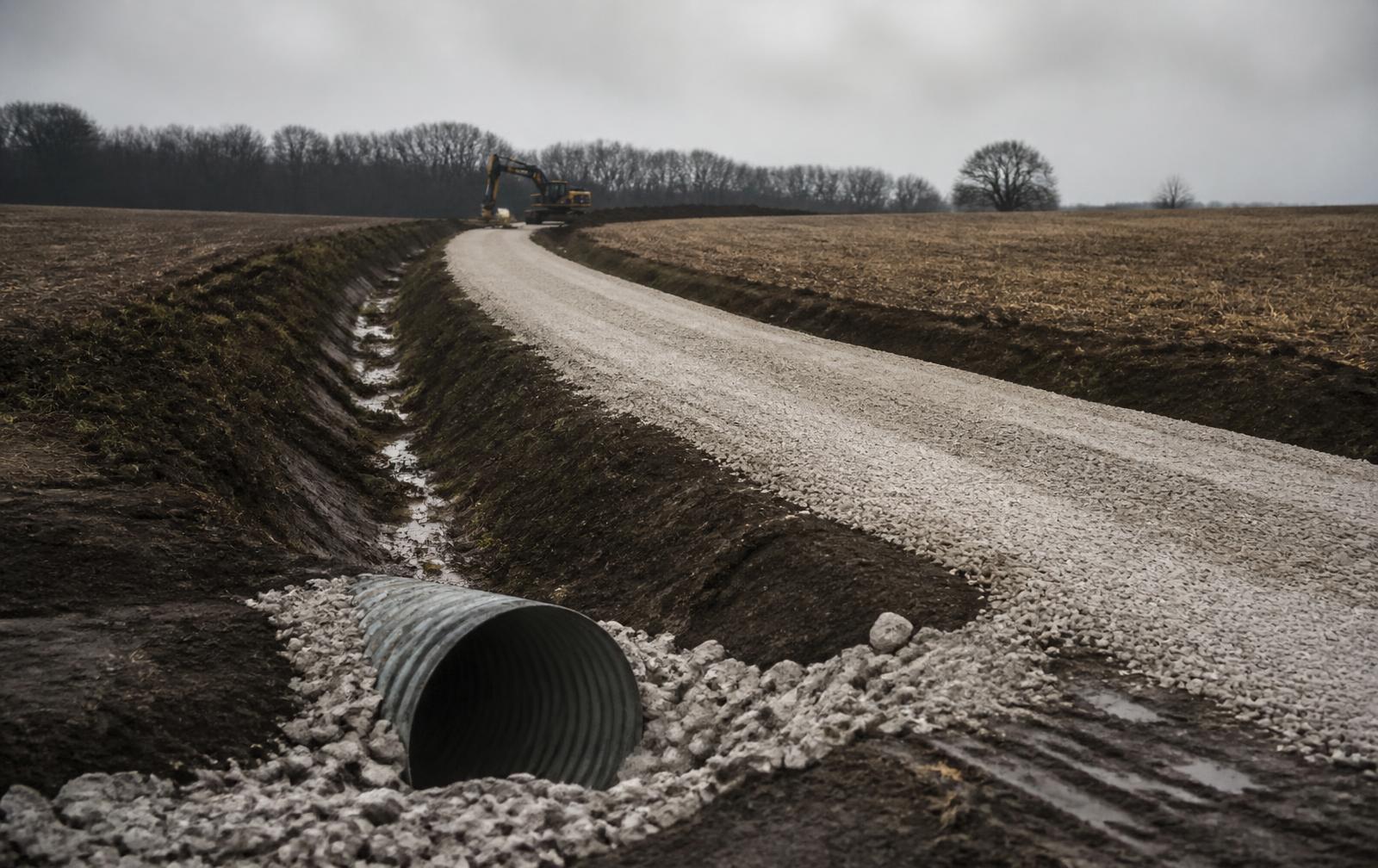 New crushed limestone rural driveway with a crowned surface and a cut drainage ditch running toward a treeline on Indiana farmland
